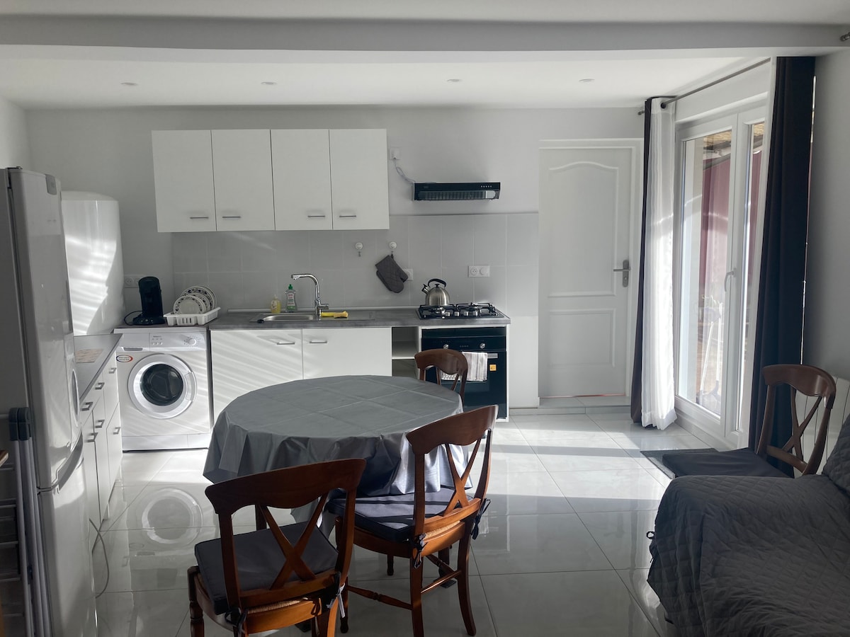 A modern kitchen area is shown, featuring white cabinetry and a gray countertop. A round dining table with wooden chairs occupies the center, while appliances like a refrigerator and washing machine are visible. Light streams in from the sliding door leading to the outdoor space.