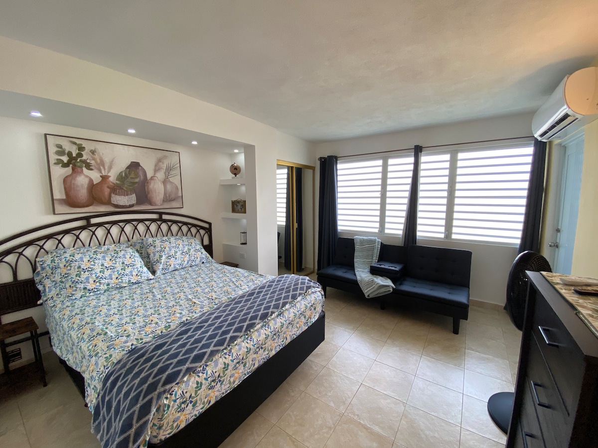A spacious bedroom features a comfortable bed with a floral-patterned duvet and coordinating throw pillows. Natural light filters through the slatted window coverings, illuminating the room's neutral color palette. A seating area with a dark sofa provides additional space for relaxation.