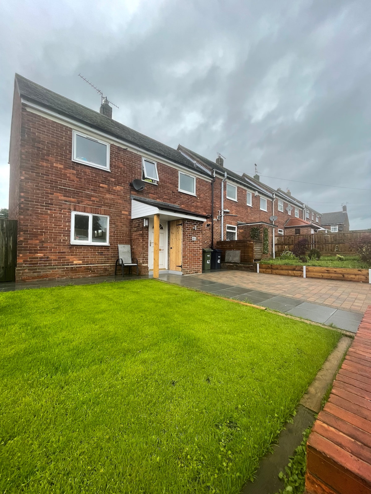 A semi-detached brick house stands in a quiet village setting, featuring a well-manicured front garden with short grass and paving. The entrance is framed by a small porch. Nearby, residential homes are visible, and gray clouds create a soft diffused lighting.