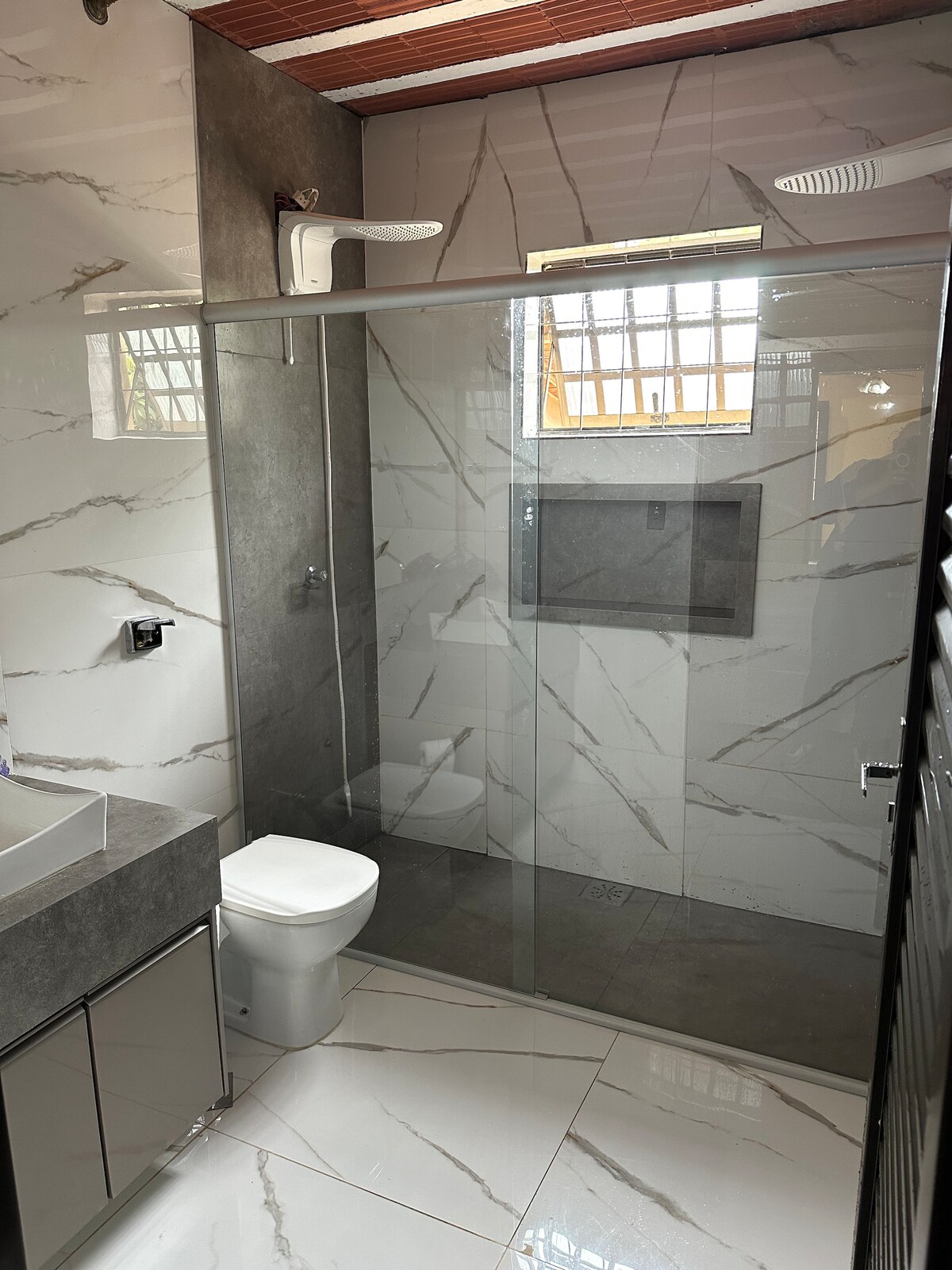 A modern bathroom features a glass-enclosed shower area with marble-patterned walls. A sleek toilet is positioned to one side, and a vanity with a basin is visible with smooth surfaces. Natural light enters through a window adorned with metal grilles.