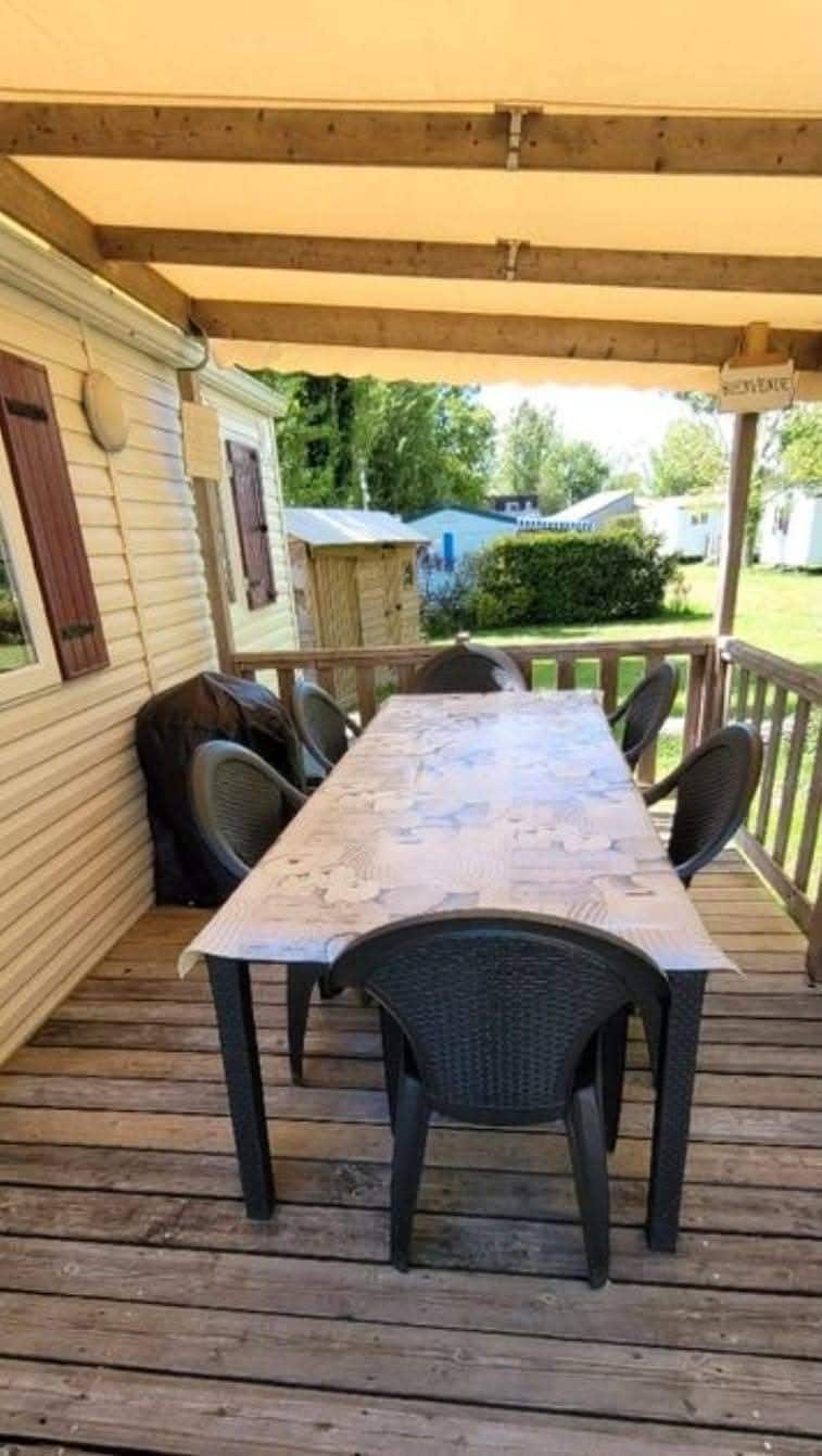 A spacious wooden terrace is featured, equipped with a large table surrounded by six dark wicker chairs. The area is covered by a roof, providing shade. Nearby greenery can be seen beyond the railing, creating a pleasant outdoor dining environment.