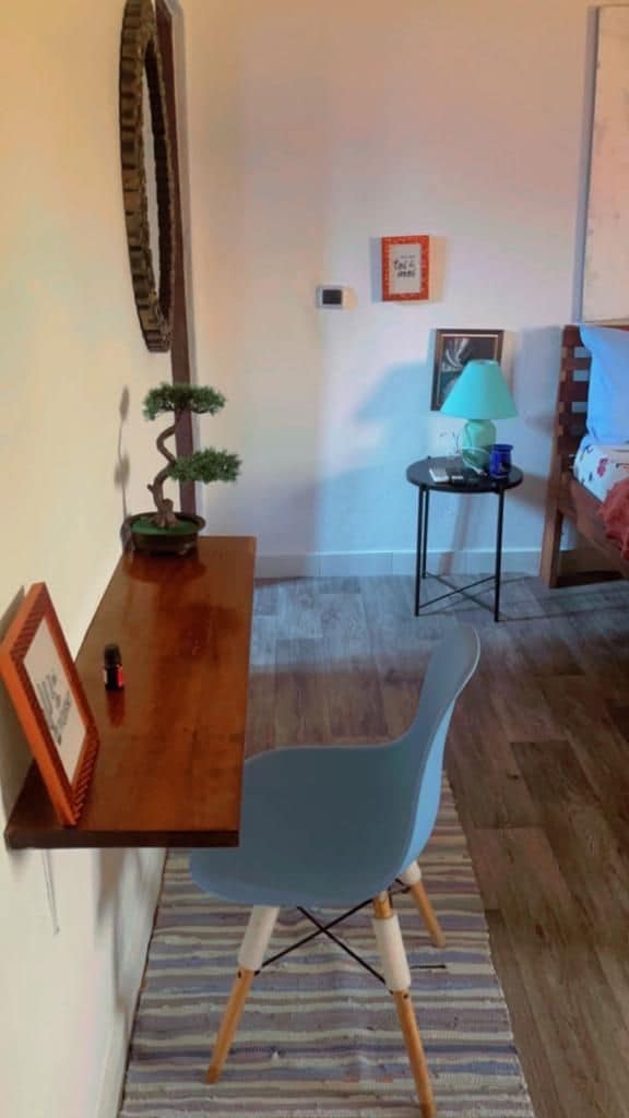 A small desk made of warm wood faces a light blue chair. A decorative bonsai plant is positioned to the left, with framed artwork placed nearby. The space features a striped area rug, while a bedside table with a green lamp and small decorative items is visible in the background.