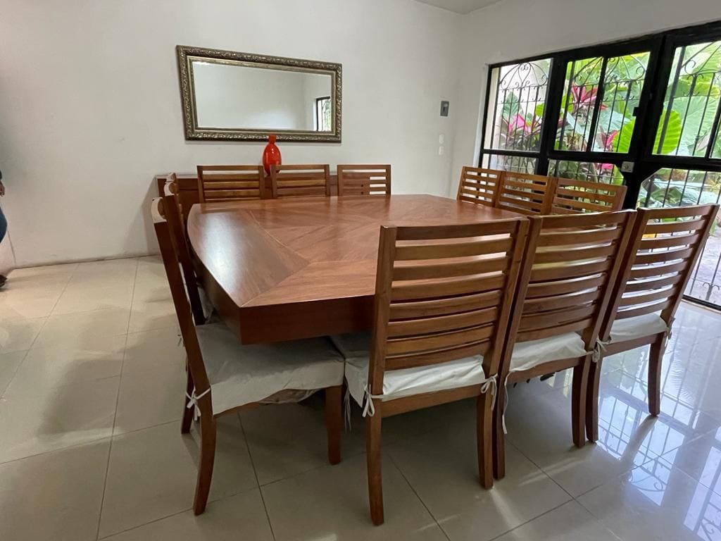 A large dining table with a polished wooden surface is surrounded by multiple wooden chairs, featuring padded seats. A decorative orange vase is placed at the center of the table. Natural light filters in through large windows adorned with greenery, creating an open and inviting setting.