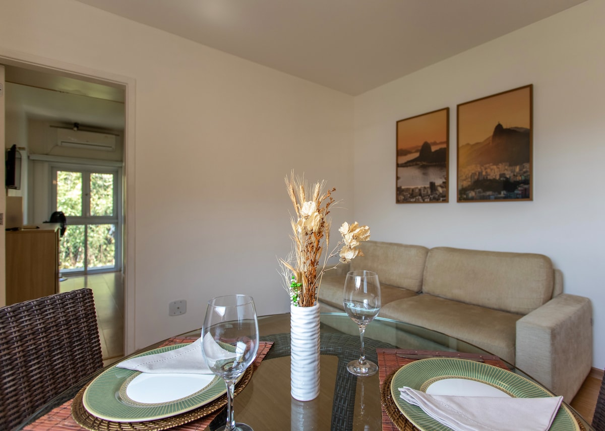 A light-filled living area features a glass dining table set for two, complemented by woven placemats. A beige sofa occupies one side, with two framed pictures of a scenic view adorning the light-colored walls, providing a homely touch.