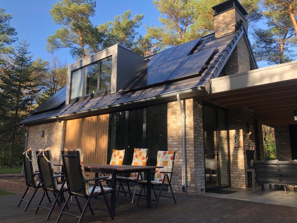 A modern house featuring a large terrace with a dining table and seating for several guests. Sunlight filters through the trees, highlighting the brick exterior and sleek design. Glass doors provide a view into the interior, while solar panels are positioned on the sloped roof.