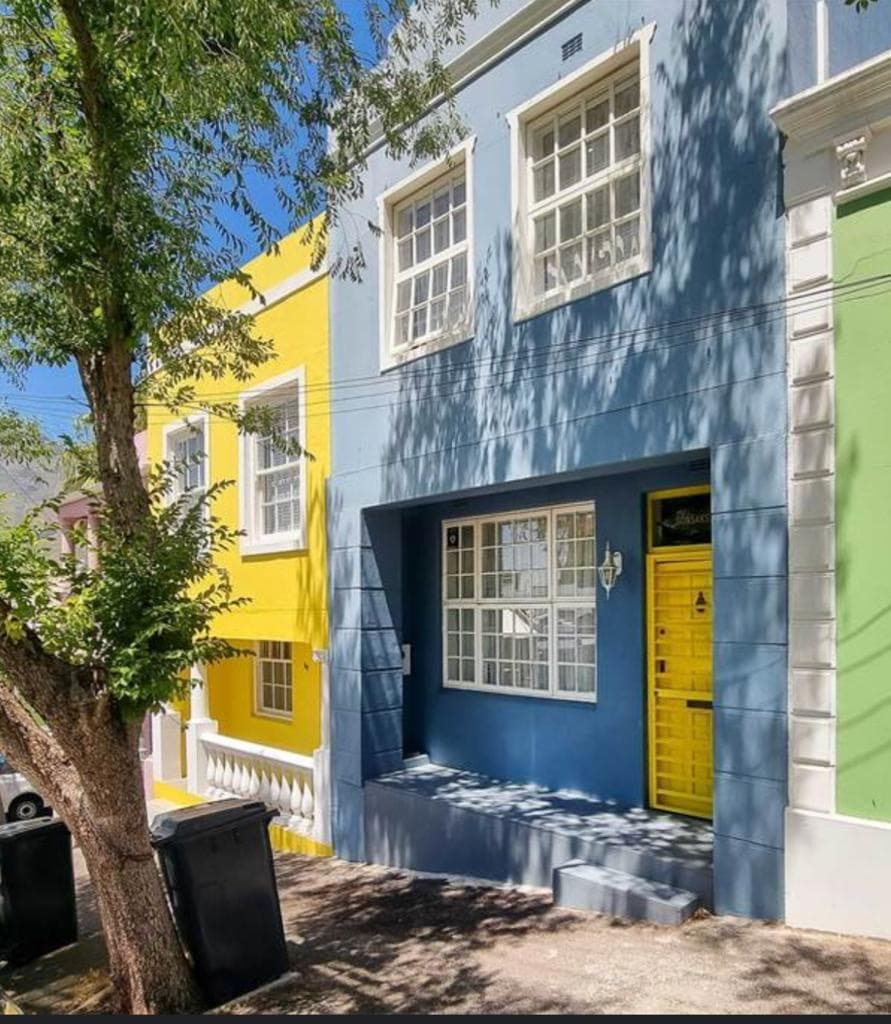 A charming row of houses in the Bo-Kaap neighborhood, featuring vibrant blue and yellow facades. The house in the center has large windows that allow natural light in, while a bright yellow door stands out. Lush green trees provide shade along the street.