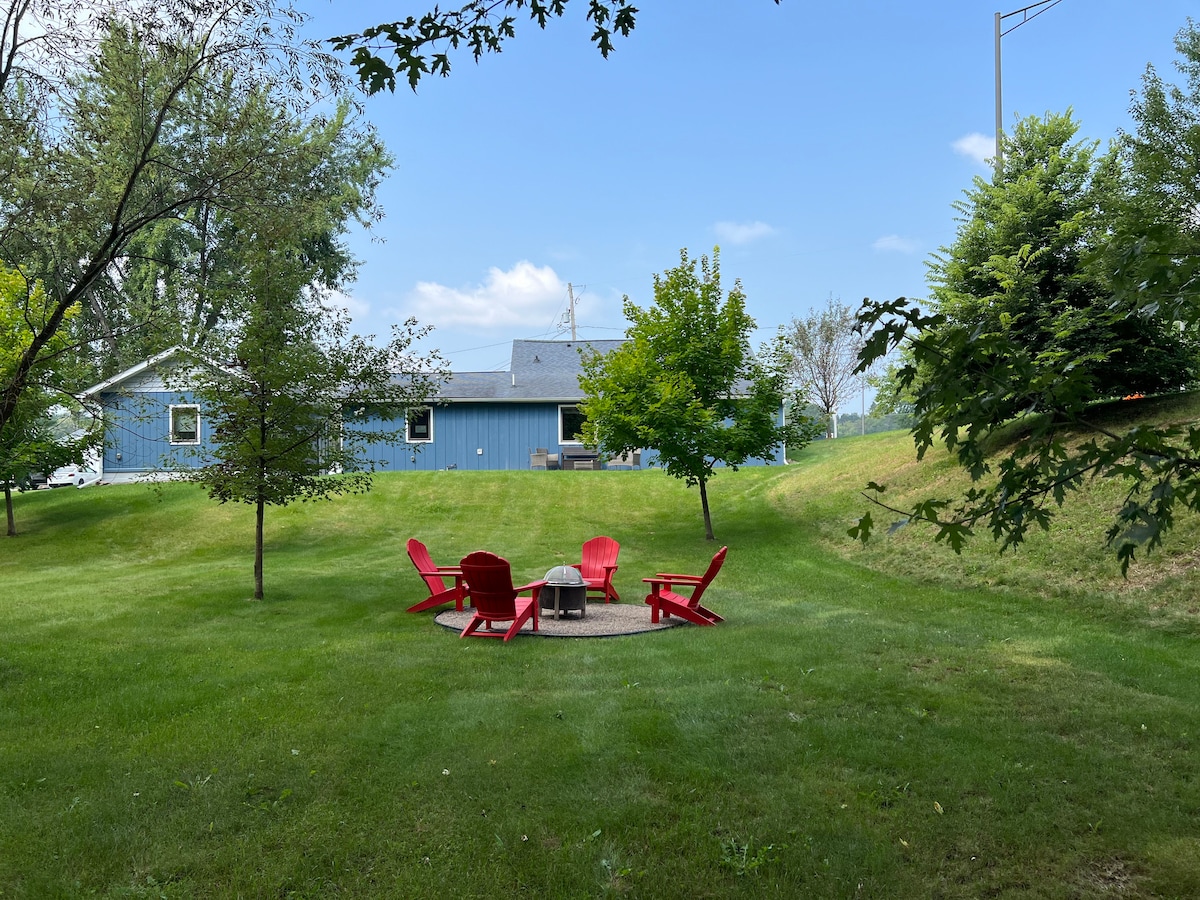 A serene outdoor space features a circular arrangement of four red chairs surrounding a fire pit. Lush green grass and trees frame the area, while a blue home stands in the background under a clear sky, providing a tranquil setting for relaxation.