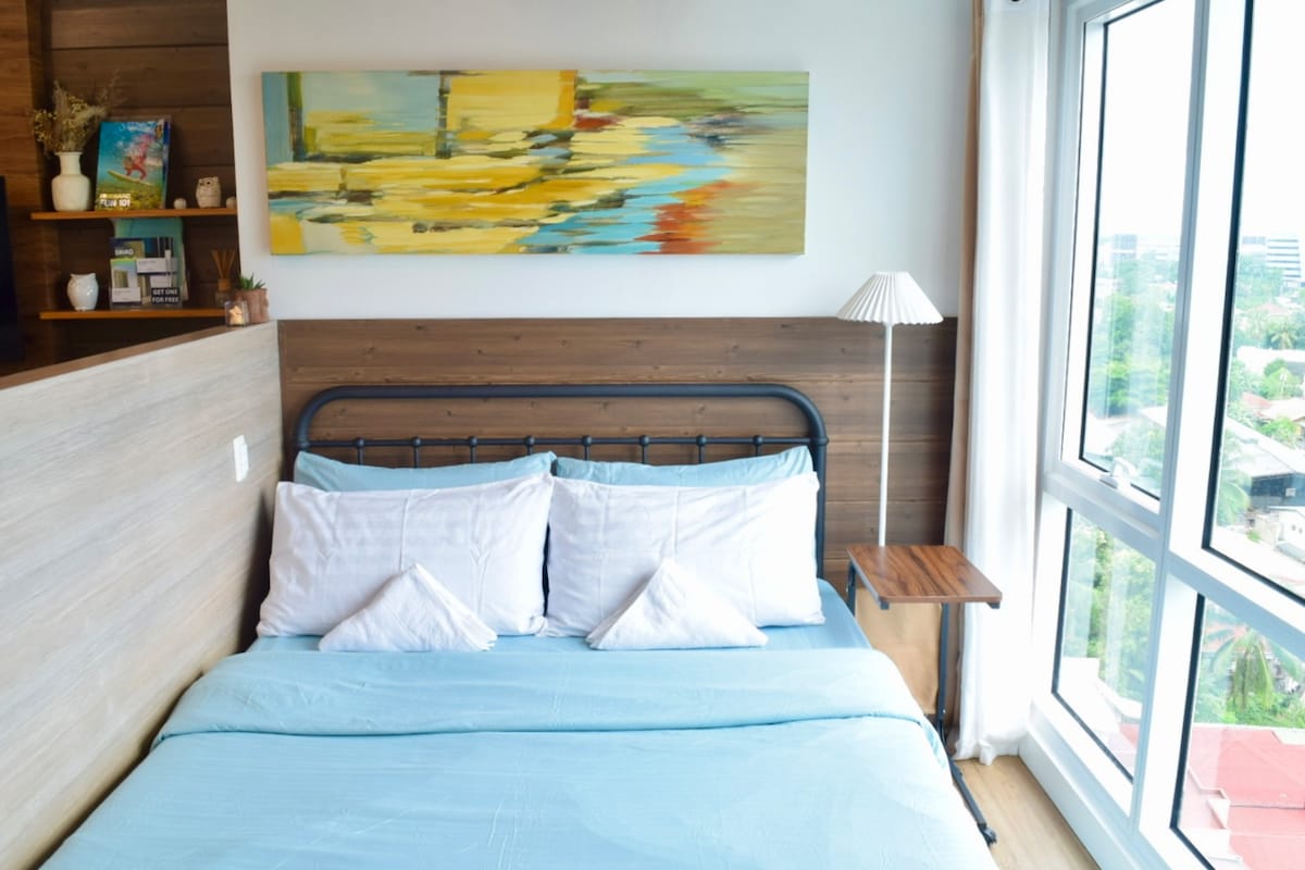 A serene sleeping area is featured, showcasing a queen-sized bed adorned with a light blue bedding set and white pillows. A modern wall-mounted lamp is positioned beside a wooden side table, while a colorful abstract painting hangs above the bed, adding a touch of artistry to the space.