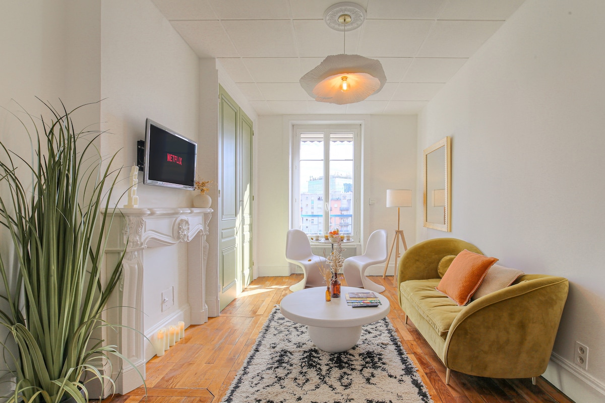 A bright living area features a cozy green sofa and modern white chairs surrounding a circular coffee table. A decorative plant adds a touch of greenery, while a television mounted on the wall offers entertainment. Natural light floods in through the window, enhancing the space.