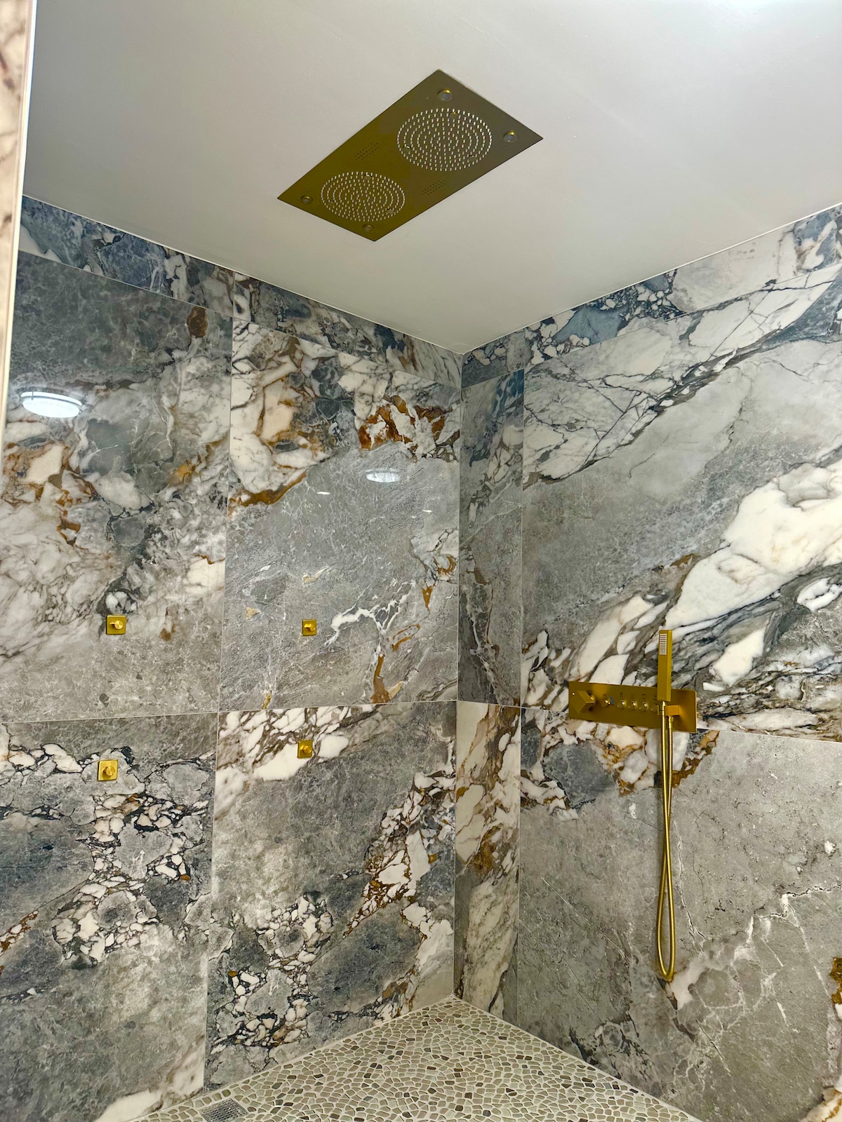 The shower space is enveloped in elegant marble tiles with a mix of grey tones and gold veining. A sleek golden showerhead is mounted on the wall, complemented by matching hardware. The floor features small, smooth pebbles, offering a natural texture.