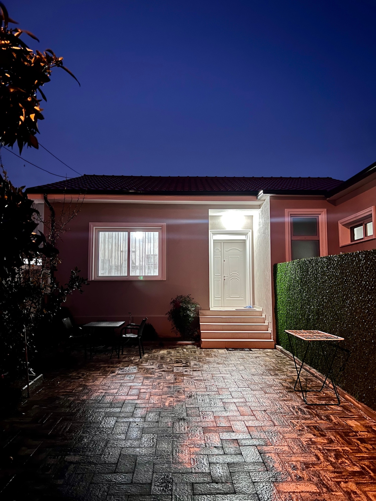 A contemporary exterior view of the accommodation is presented at twilight. A welcoming entrance with a light-colored door is flanked by a well-lit pathway and a small sitting area. Lush greenery lines the space, enhancing the inviting atmosphere.