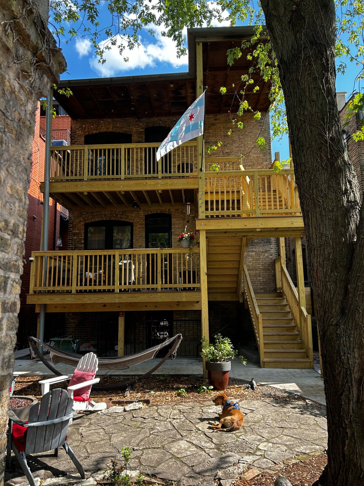 An exterior view of a three-story brick building featuring wooden decks with railings and steps leading to the upper levels. A Chicago flag is displayed prominently. The shaded yard contains a hammock, stone patio, and seating area, with a friendly dog resting nearby.