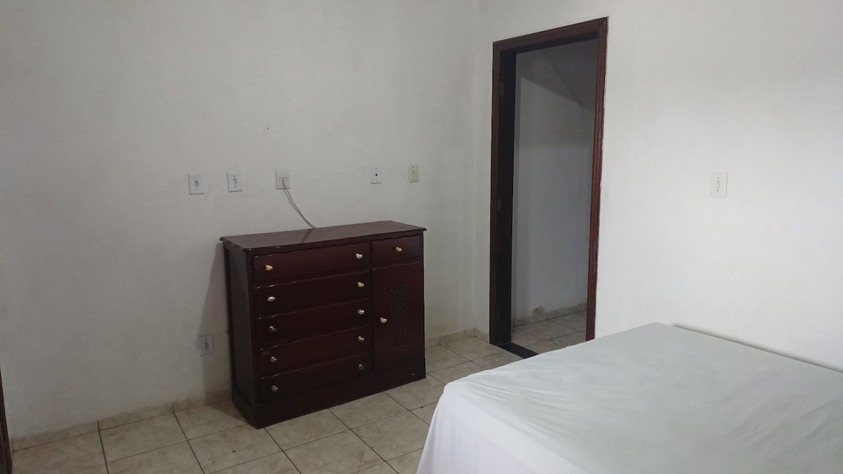 An empty room features a beige tiled floor and plain white walls. A dark wooden dresser with six drawers is positioned against one wall. An open door leads to another space, providing access to the adjacent area.