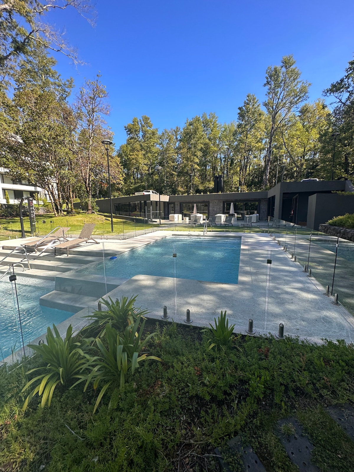 An outdoor area features a modern infinity pool surrounded by a glass barrier. Lounge chairs are positioned on the pool deck. The lush greenery of trees and plants create a serene backdrop, while a contemporary building provides seating and dining options in the distance.