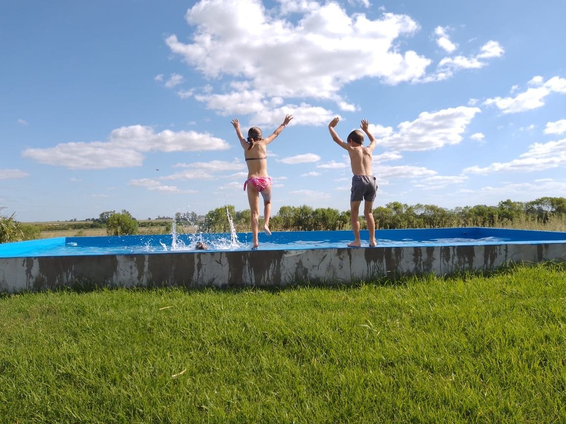 Two children are shown mid-air as they jump into a refreshing blue pool surrounded by lush green grass. The sky is partly cloudy, with fluffy white clouds against a backdrop of blue. The scene captures a moment of joy and playfulness in a natural setting.