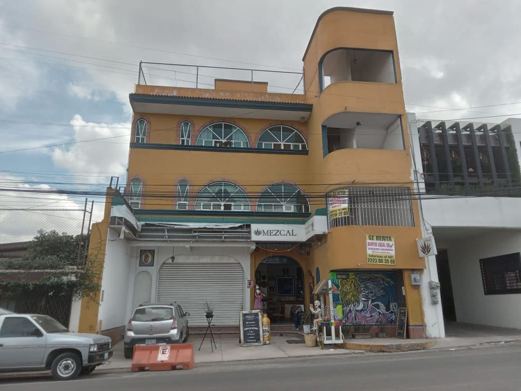 The exterior of a three-story building features arched windows and a warm yellow facade. A sign indicating a mezcal establishment is visible, while parked vehicles line the street in front. An outdoor seating area can be seen beneath the overhang.