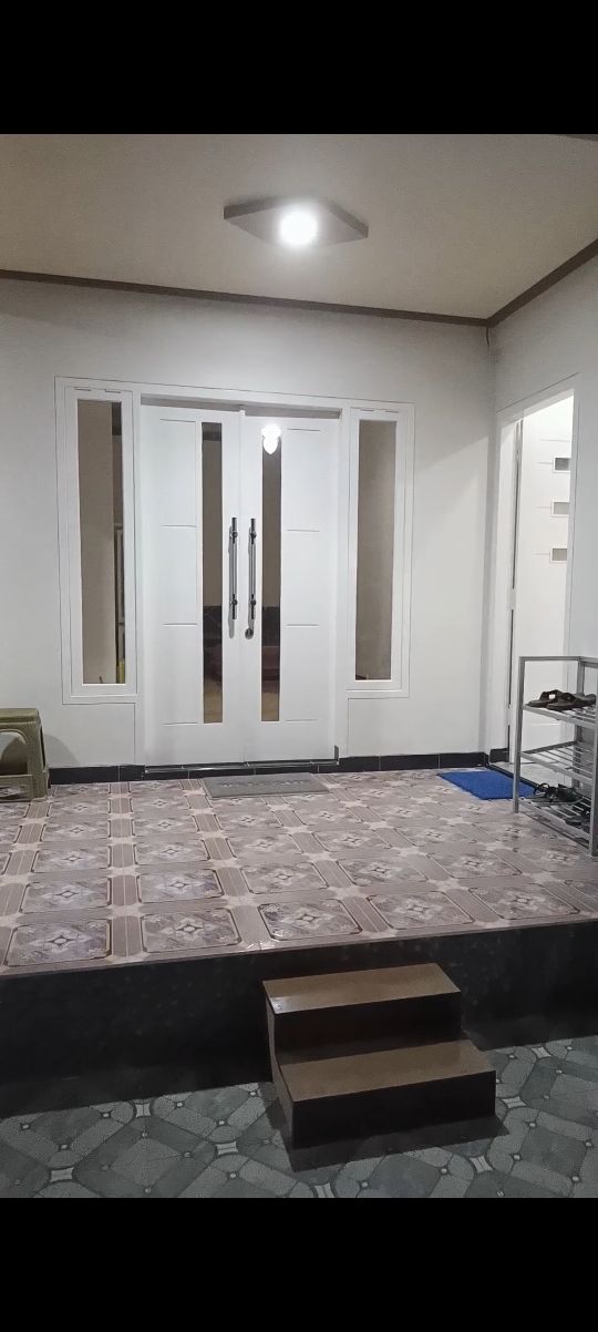 The entrance features a set of modern double doors framed by large windows. A patterned tile floor extends across the entryway, complemented by a small set of wooden steps leading up to the main area. A neatly placed doormat adds to the welcoming space.