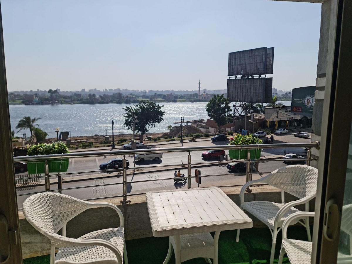 A balcony offering a view of the Nile River features a small white table and two matching chairs, positioned against a backdrop of palm trees and city buildings. The scene includes a clear sky, with distant structures visible along the riverbank.