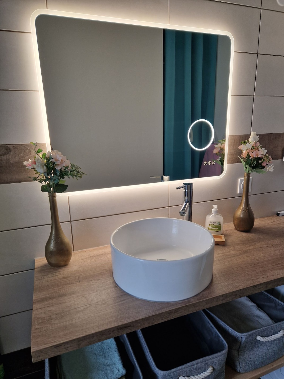 A modern bathroom sink area is displayed, featuring a circular white basin on a wooden countertop. Two small vases with flowers accent the space. A backlit mirror above adds warmth, while organized storage baskets are positioned below the countertop.