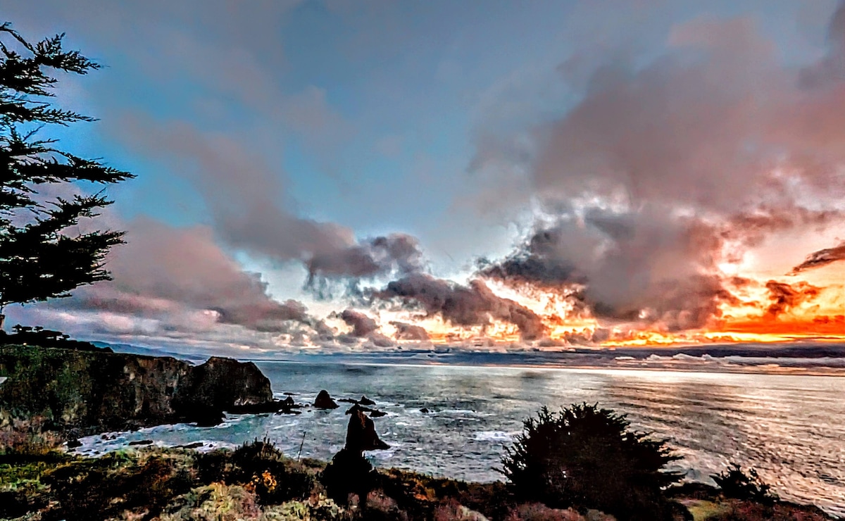 A sweeping view of the ocean is presented, framed by coastal cliffs and silhouettes of trees. The sky is filled with clouds illuminated by a warm sunset, transitioning from pale pink to vibrant orange, contrasting with the calm, reflective sea below.