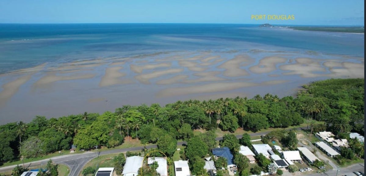 An aerial view captures the coastline, showcasing the Coral Sea and the unique patterns of the exposed seabed at low tide. Lush greenery borders the shoreline, while the structures of the neighborhood are visible, emphasizing proximity to nature and the beach.