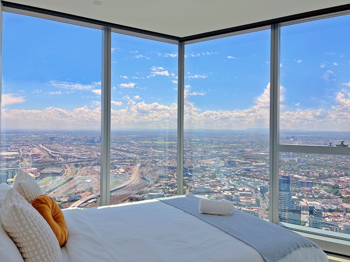 A spacious bedroom offers floor-to-ceiling windows, providing panoramic views of Melbourne's skyline and beyond. A soft blanket is neatly folded at the foot of a large bed, while a cityscape extends in the background under a bright blue sky.
