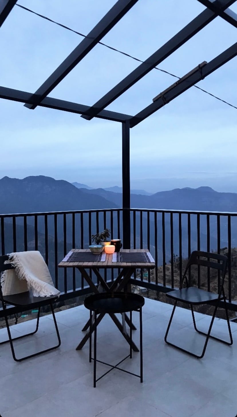 A balcony with a metal frame features a table and two chairs, providing a space to enjoy the surrounding mountain views. A gentle light from a candle on the table adds a soft glow against the backdrop of the evening sky.