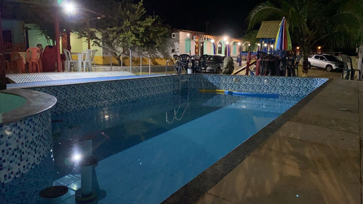 A night view of a swimming pool features illuminated water reflecting soft lights. The surrounding area includes colorful buildings and outdoor seating, with a car parked nearby. Palm trees add a tropical touch to the ambiance.