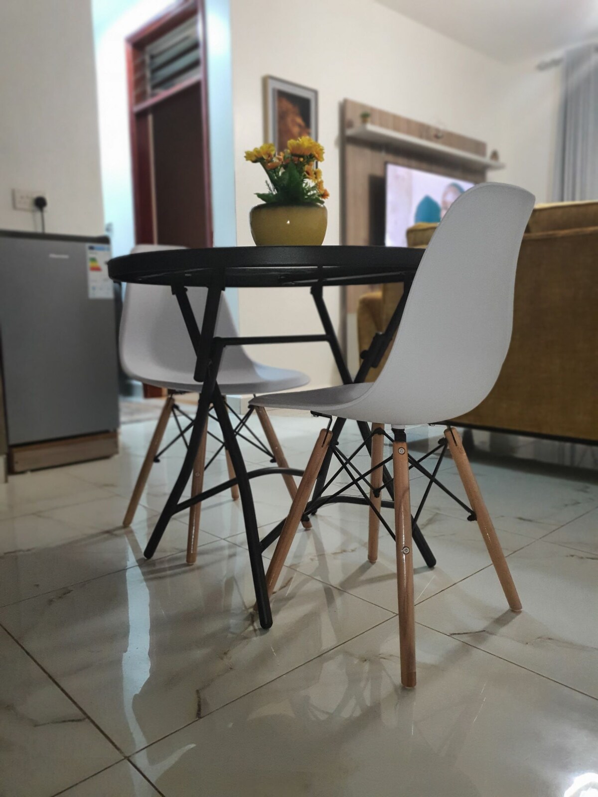 A dining area features a small round black table surrounded by two white chairs with wooden legs. A potted plant with yellow flowers is placed in the center of the table. The shiny tiled floor reflects the seating arrangement, contributing to a clean and modern aesthetic.