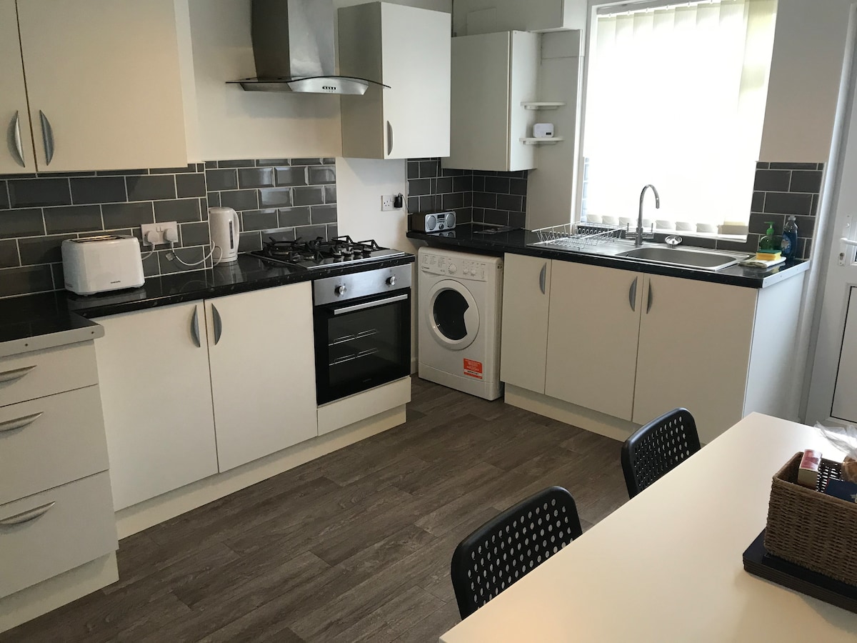 The kitchen-dining area is equipped with modern cabinetry and includes essential appliances such as a gas stove, oven, and washing machine. A clear countertop provides workspace, and a dining table is set with black chairs. Natural light enters through a window with vertical blinds.