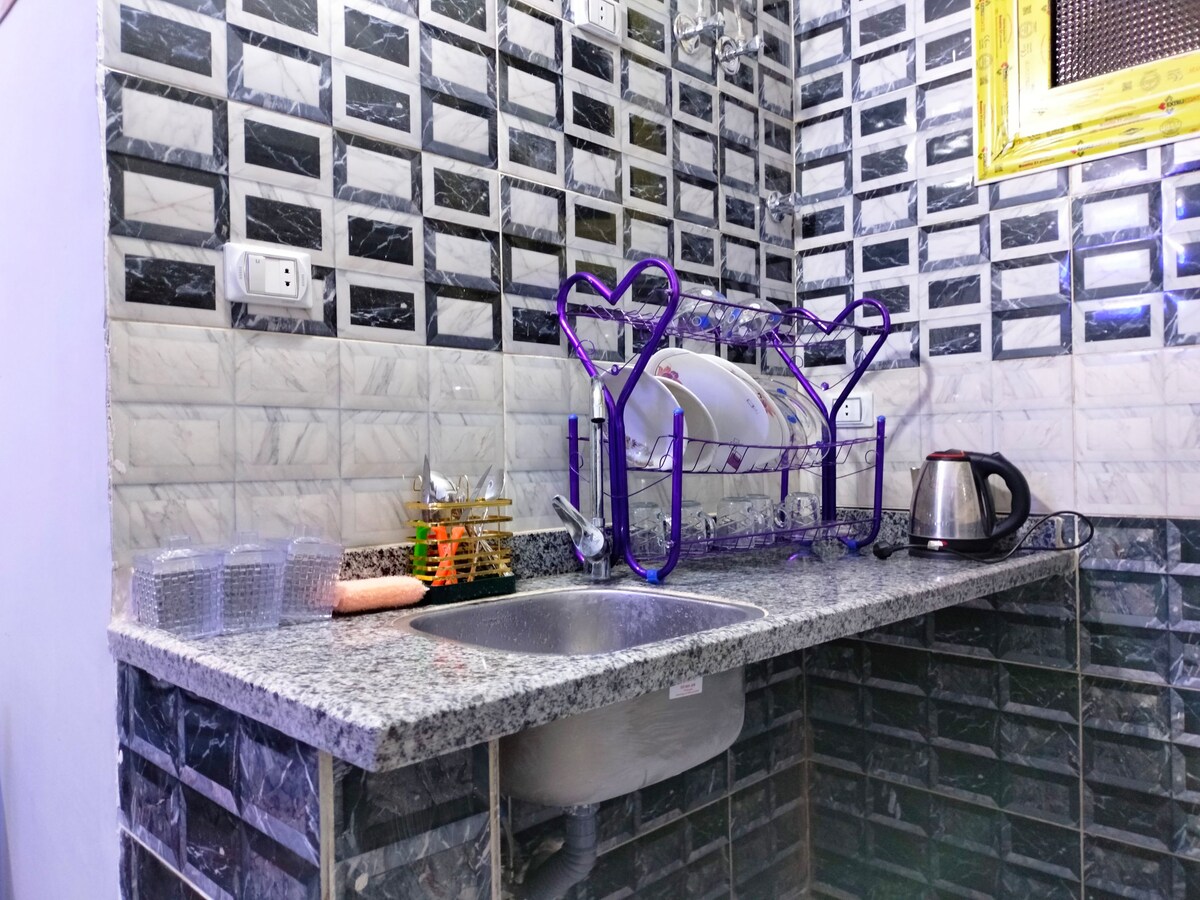 A compact kitchen area features granite countertops with a silver kettle and dish drying rack. Glass cups are organized alongside utensils. The backsplash displays a modern pattern of black and white tiles, contributing to the stylish decor.