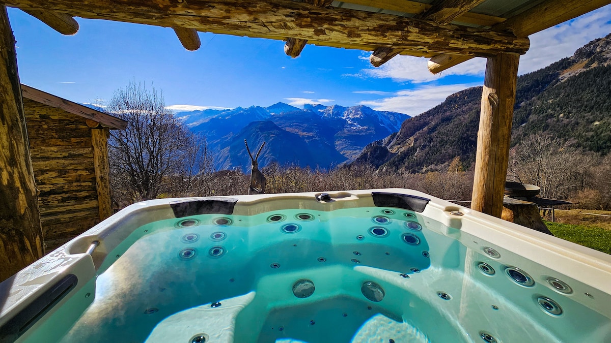 An outdoor jacuzzi is surrounded by wooden beams, offering a view of the mountains and valleys in the distance. Clear blue skies and a few clouds enhance the landscape, creating a serene setting for relaxation.