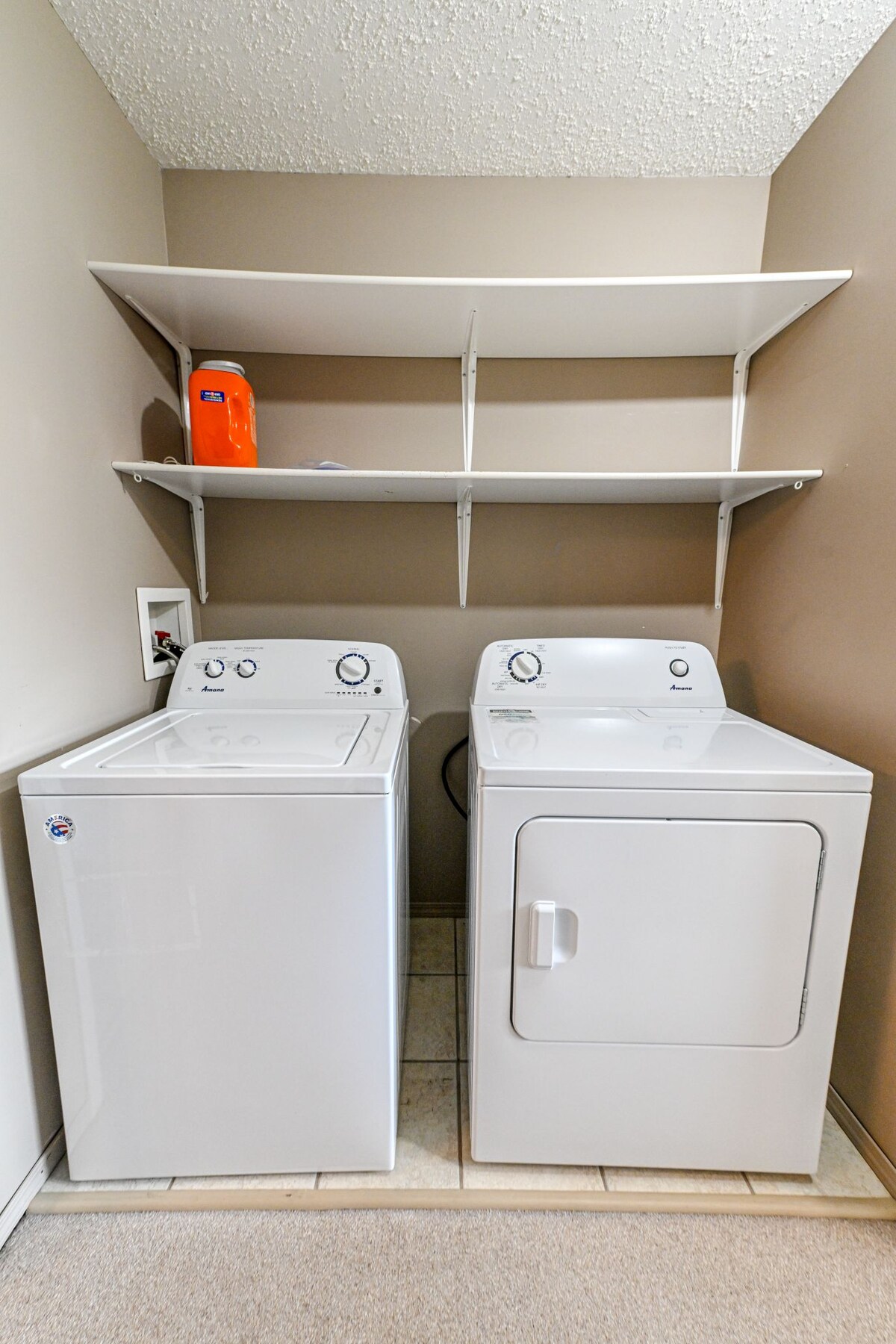 A clean laundry area is equipped with a washing machine and dryer, both in white. Above, two shelves provide storage space, with an orange bottle placed on the top shelf. The tiled floor complements the neutral wall color.