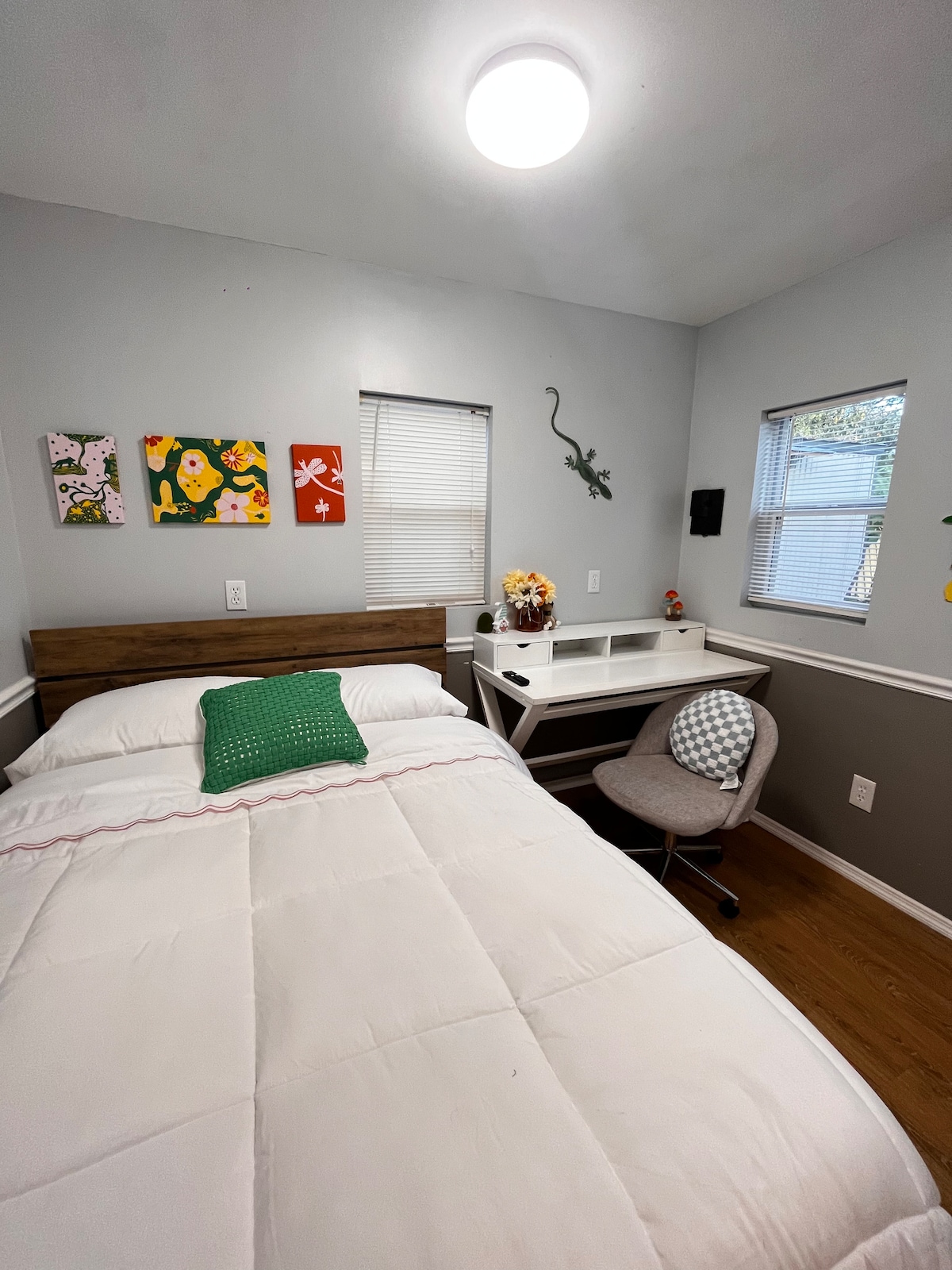 A cozy bedroom features a spacious bed with a white comforter and a green accent pillow. Two windows allow natural light to brighten the space, while a small desk and chair are positioned against the wall. Colorful artwork adds a vibrant touch to the room.