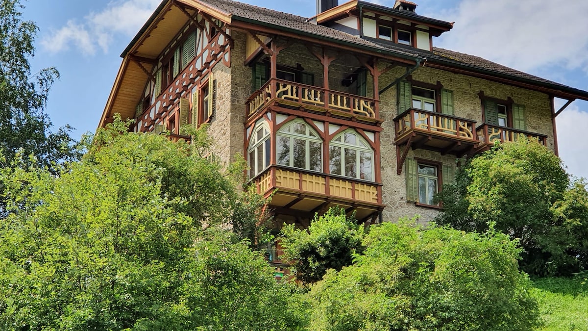 A three-story house is surrounded by lush greenery, showcasing a mix of stone and wood architecture. Large arched windows provide natural light, and balconies with wooden railings extend out from the second and third floors, offering views of the landscape.