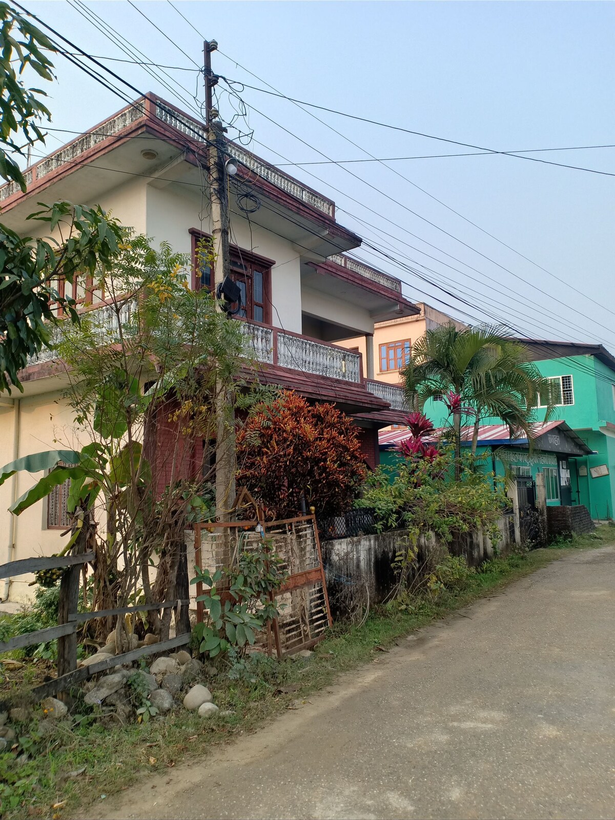 A multi-story residence is visible with a blend of architectural styles and colors, featuring greenery and blooming plants in the front yard. Power lines run overhead, and a quiet dirt road is seen leading to the entrance, providing a sense of accessibility to the property.