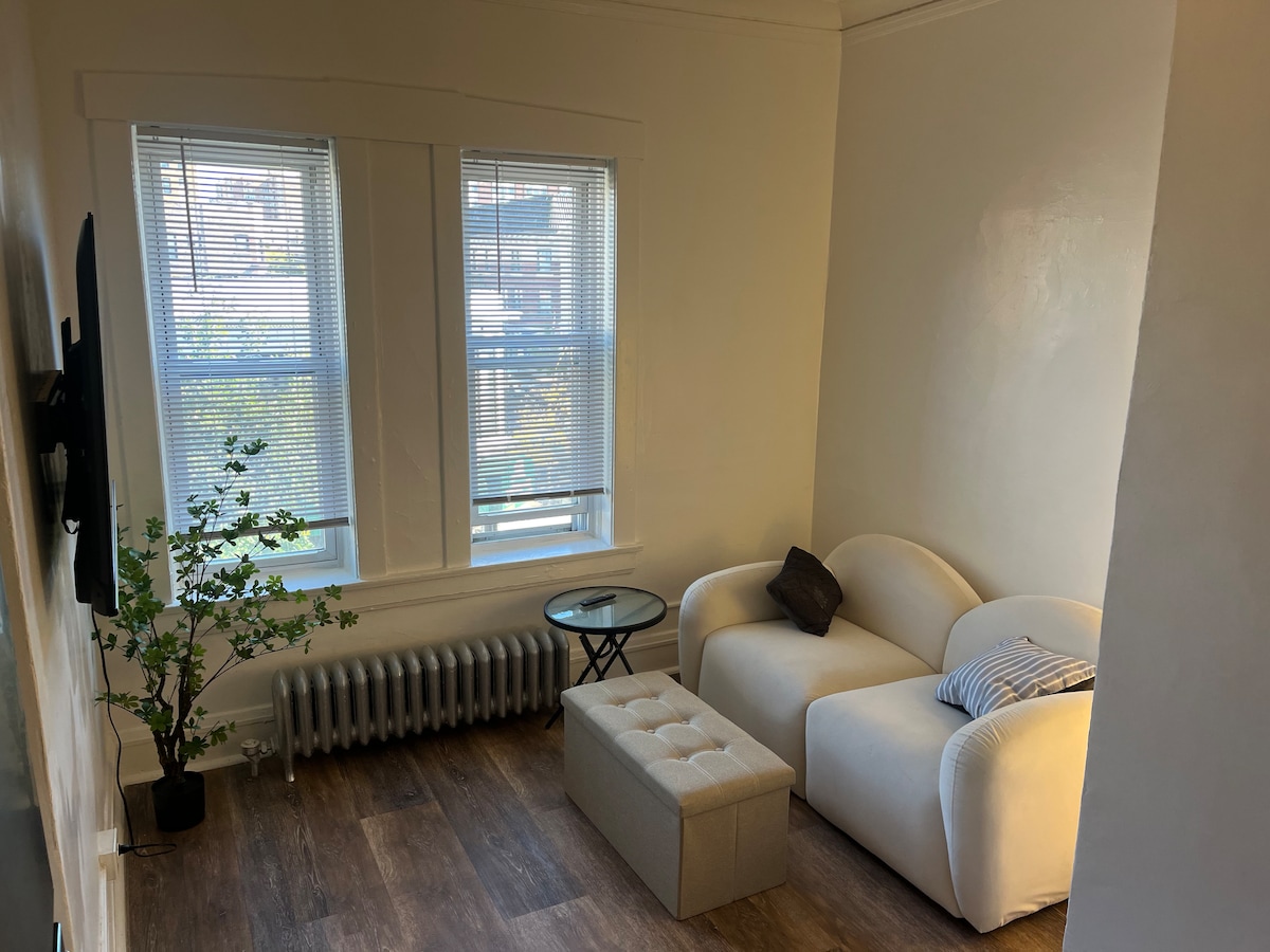 A bright living area is showcased with two windows allowing ample natural light. A cozy white couch sits beside a small round table, accompanied by a tufted ottoman. A green plant adds a touch of color, while a radiator is visible beneath the windows.