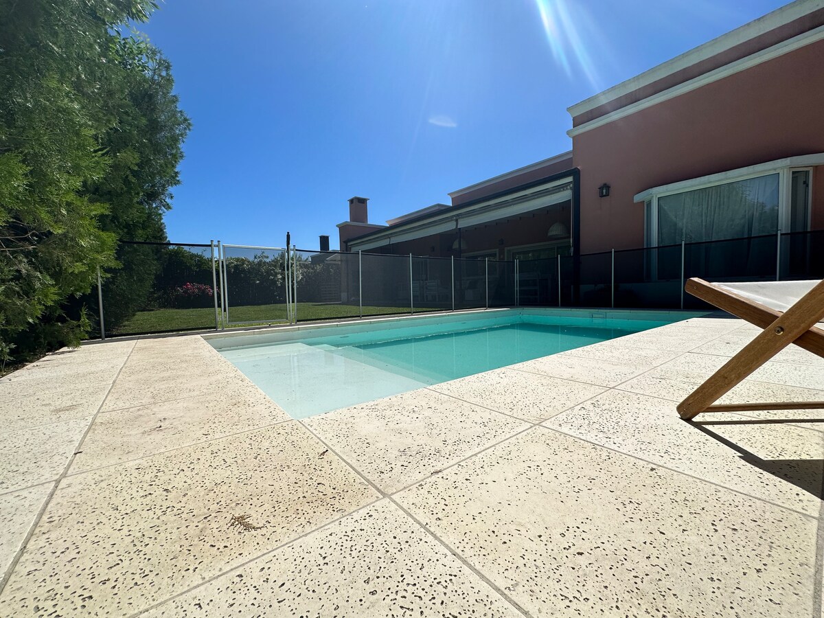 A serene swimming pool is framed by a safety fence, with clear water reflecting the sunlight. The surrounding area includes a spacious patio that features a sun lounger and grass, providing a tranquil outdoor setting.