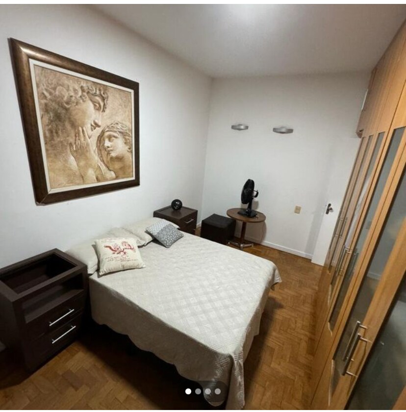 The private room features a spacious bed covered with a light, patterned blanket. A decorative artwork hangs prominently above the bed. A wooden nightstand and a cabinet with glass doors are visible, alongside a small fan placed on the floor.