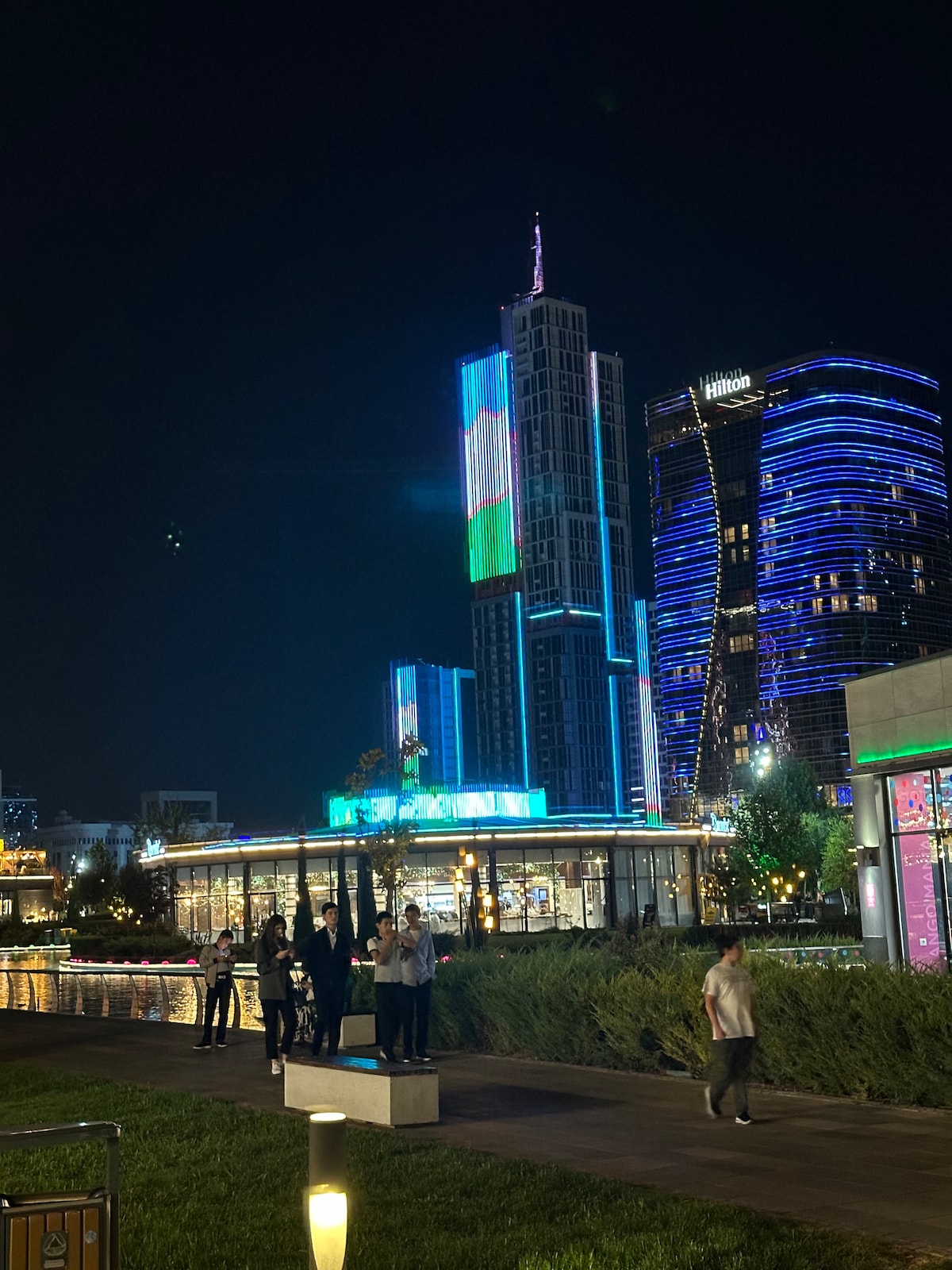 A vibrant urban scene is captured at night, featuring illuminated skyscrapers with colorful projections. The lively waterfront area showcases people gathered near a modern building, surrounded by lush greenery and soft lighting that creates a welcoming atmosphere.