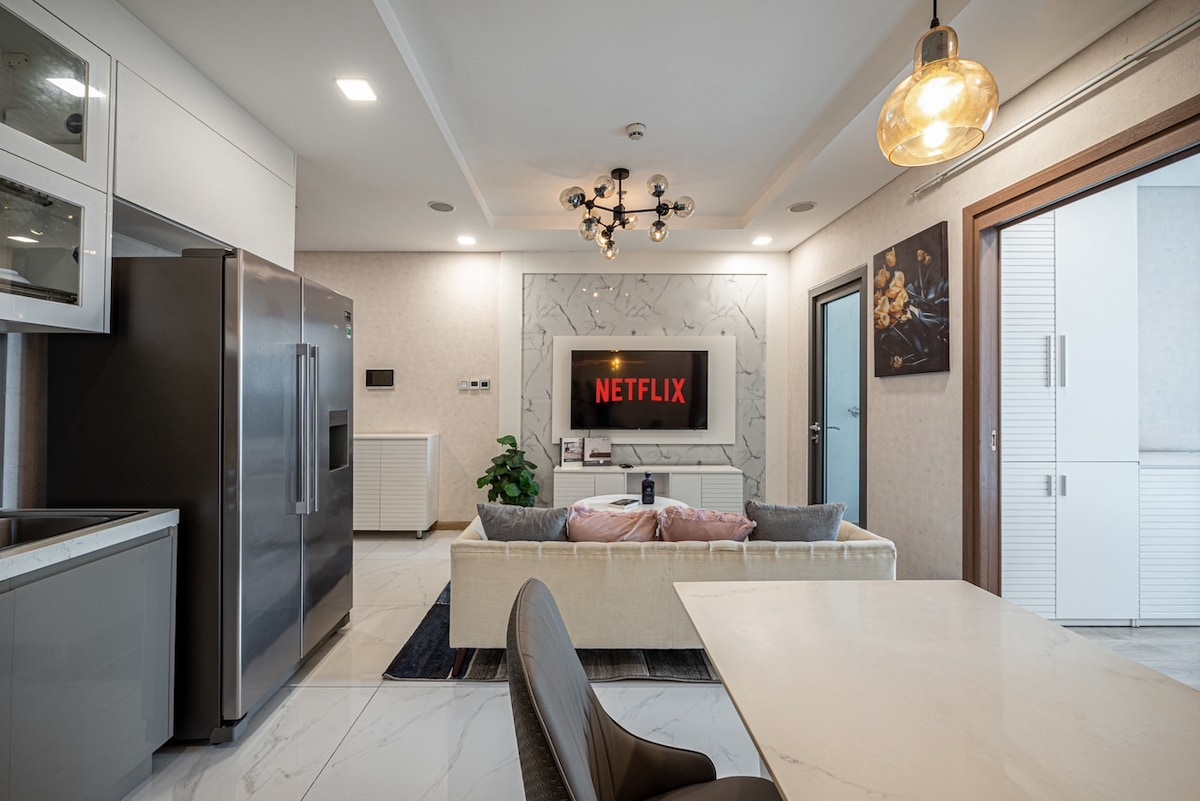A modern living space features a comfortable sofa with cushions, facing a wall-mounted television displaying Netflix. A sleek kitchen is visible in the background, complemented by a dining table and stylish lighting fixtures. Large windows bring in natural light, enhancing the open atmosphere.