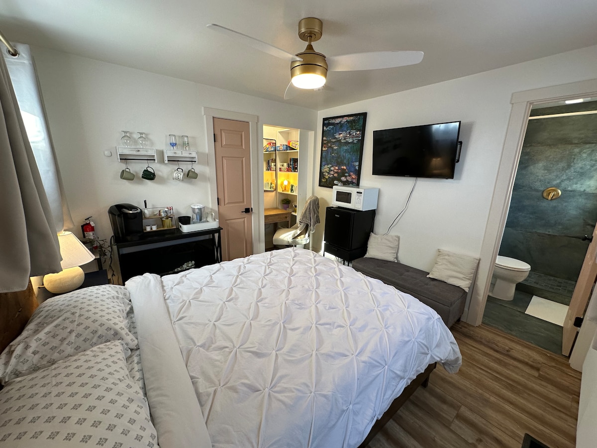 A cozy mini-suite features a queen bed with a soft blanket. A wall-mounted TV is positioned opposite. A kitchenette with coffee supplies and a fridge is alongside a small seating area. A door leads to a stylish bathroom with modern fixtures.