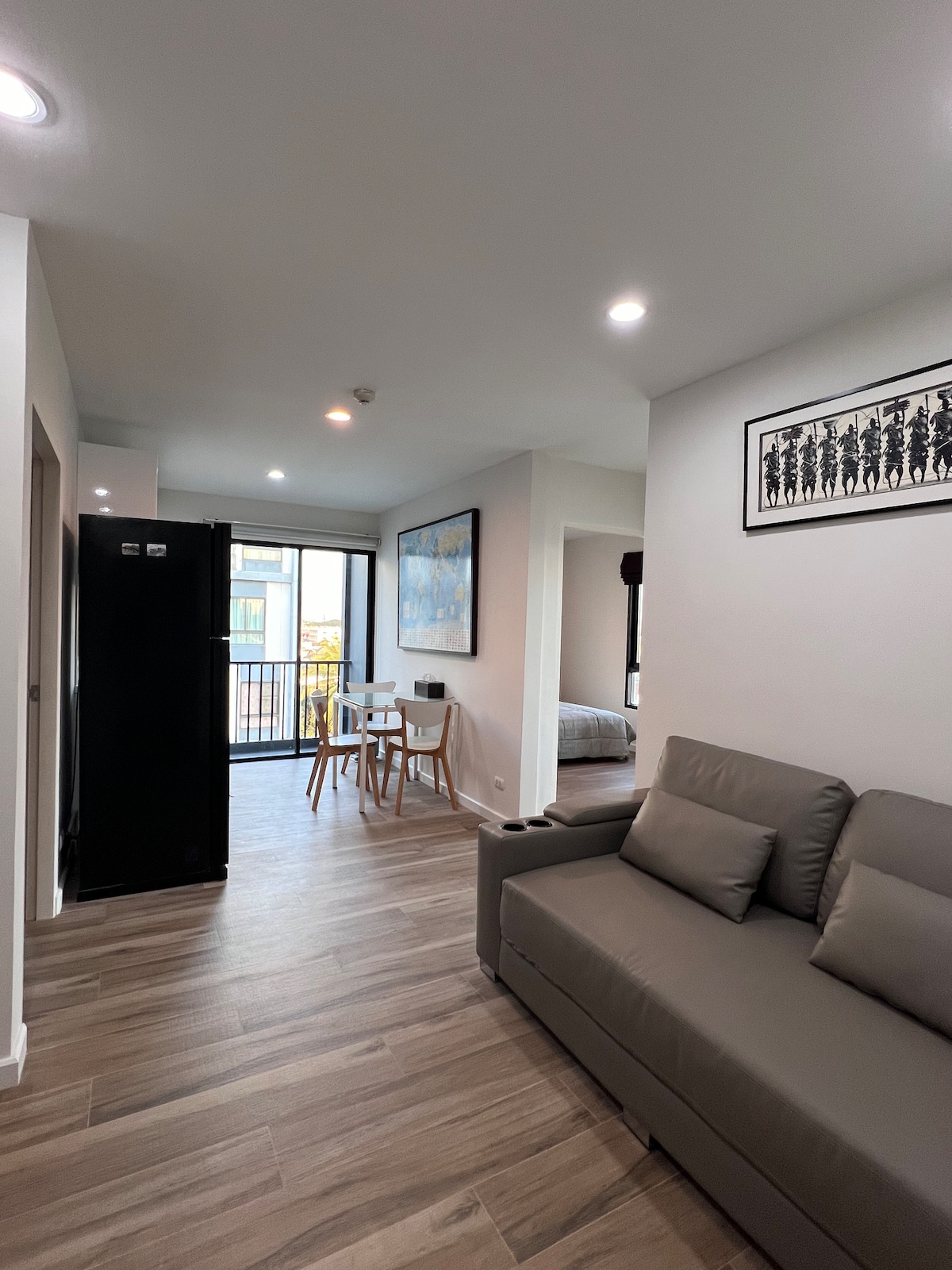 An open living area is showcased with a modern gray sofa and a dining table for two, positioned near a large sliding door that leads to a balcony. Natural light fills the space, which features light wood flooring and neutral wall colors.