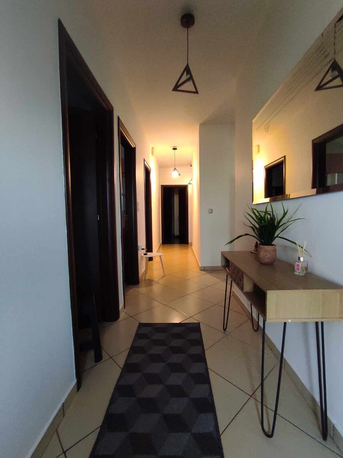 A hallway is shown, featuring a geometric patterned rug and a sleek wooden console table. Pendant lights illuminate the space, while a small potted plant adds a touch of greenery. The corridor leads to various doorways, enhancing the feeling of spaciousness.