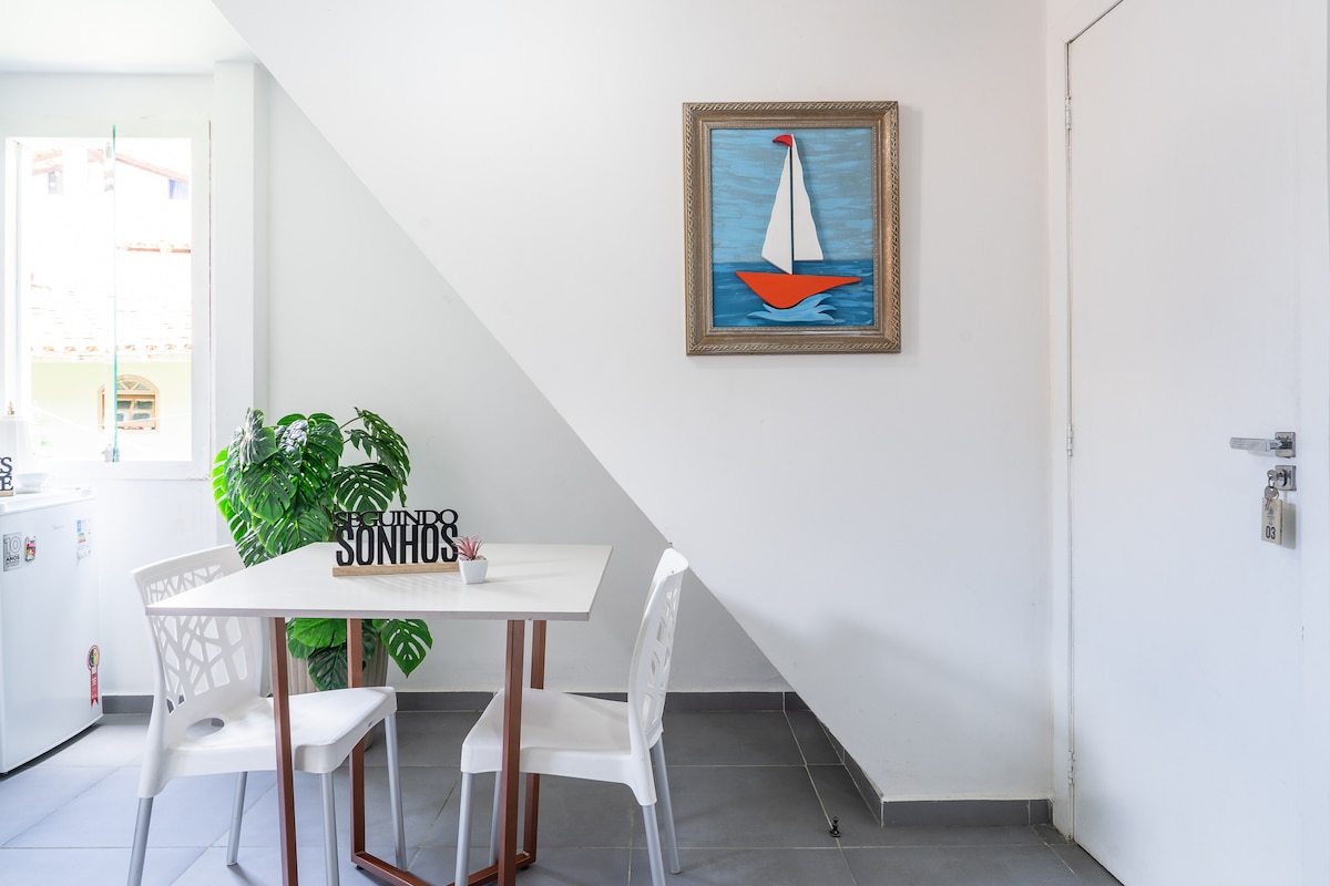 A bright dining area features a small white table surrounded by transparent chairs. A leafy green plant adds a touch of nature. A colorful painting of a sailboat is displayed on the wall, enhancing the space's light and airy feel.