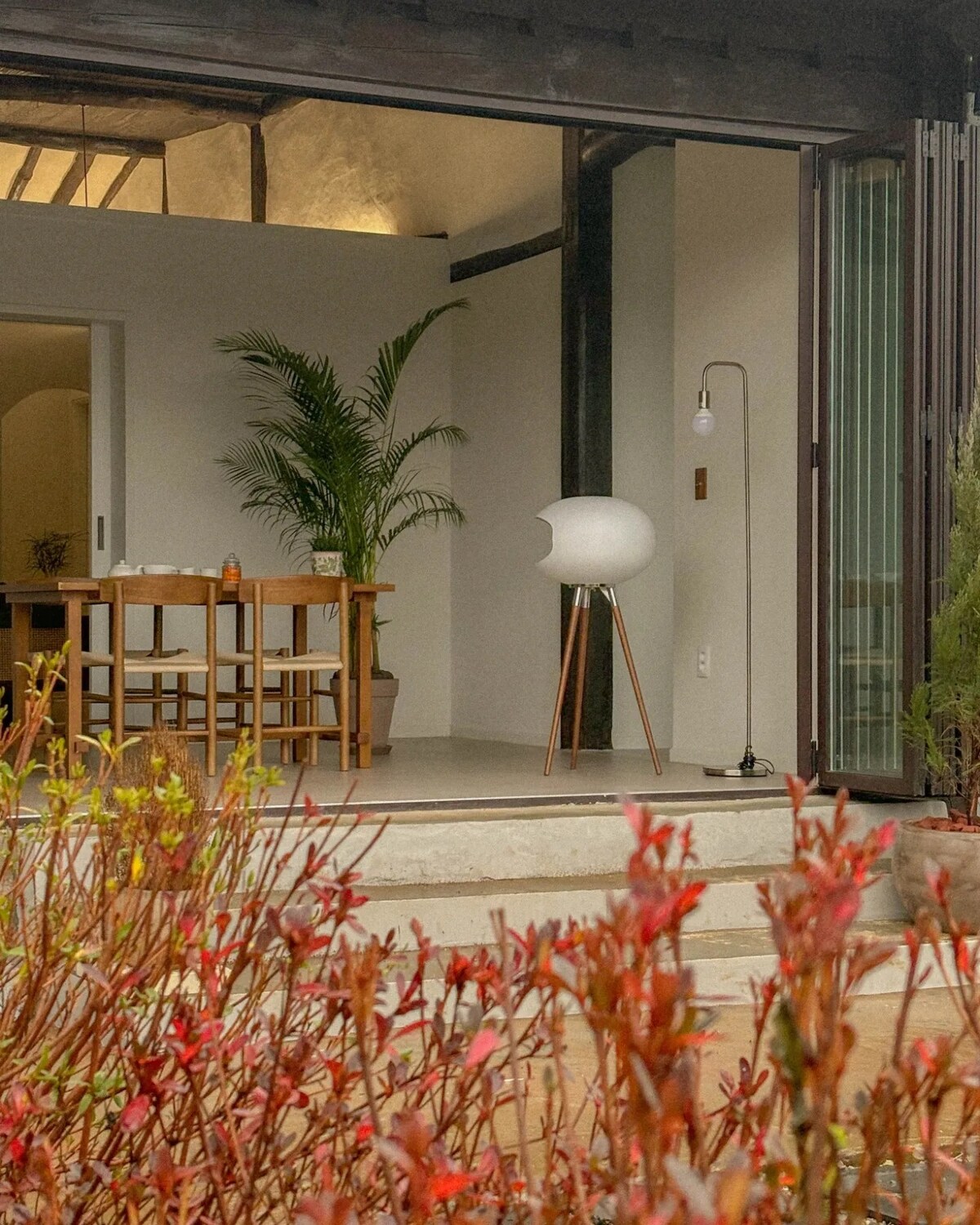 A serene outdoor view reveals a minimalistic entryway with wooden furniture, featuring a dining table and chairs. A modern lamp stands beside a sculptural element. Lush greenery and vibrant flowers frame the space, creating a connection between indoor and outdoor environments.