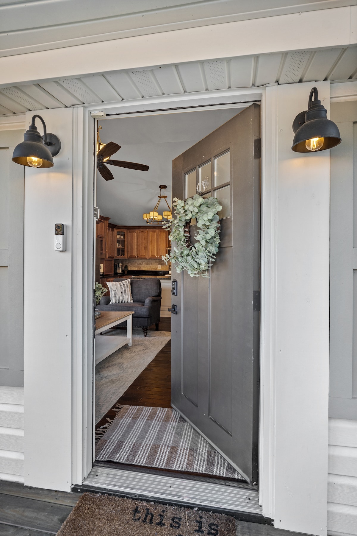 A front door is opened wide, revealing a welcoming view into a stylish interior. Light fixtures illuminate the entrance, and a decorative wreath adorns the door. A neutral rug lies at the threshold, enhancing the inviting atmosphere of the entryway.
