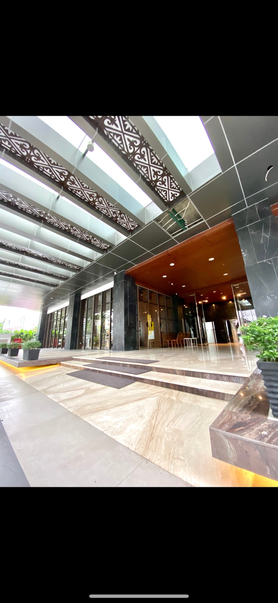 The entrance of the building is framed by sleek glass doors and large windows, allowing natural light to fill the lobby. Decorative metalwork is prominent above the entryway, while sophisticated stone flooring complements the elegant atmosphere. Potted greenery adds a touch of nature to the setting.