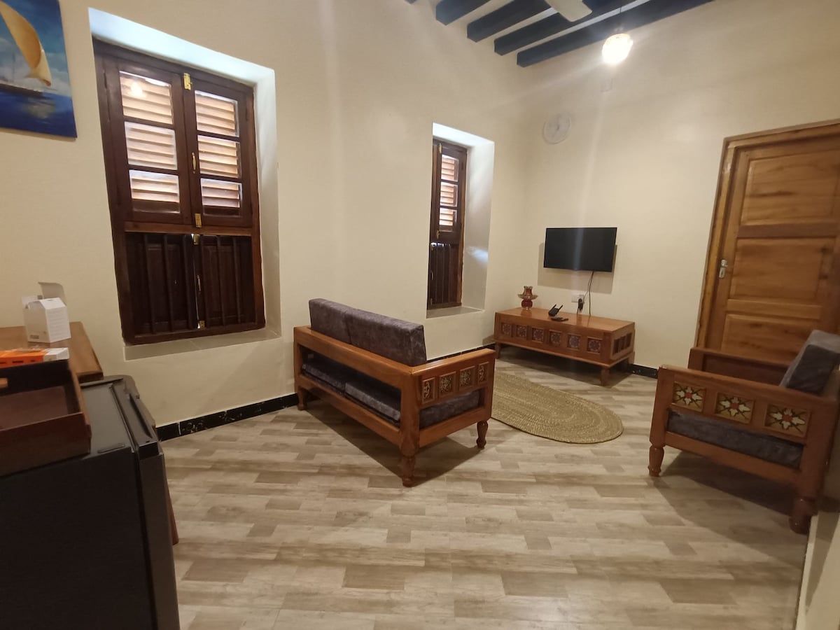 A welcoming living space features wooden furniture with intricate detailing, complemented by two chairs and a low coffee table. Natural light filters through the charming shuttered windows, and a mounted television is positioned opposite the seating area. The warm wooden door is visible in the background.