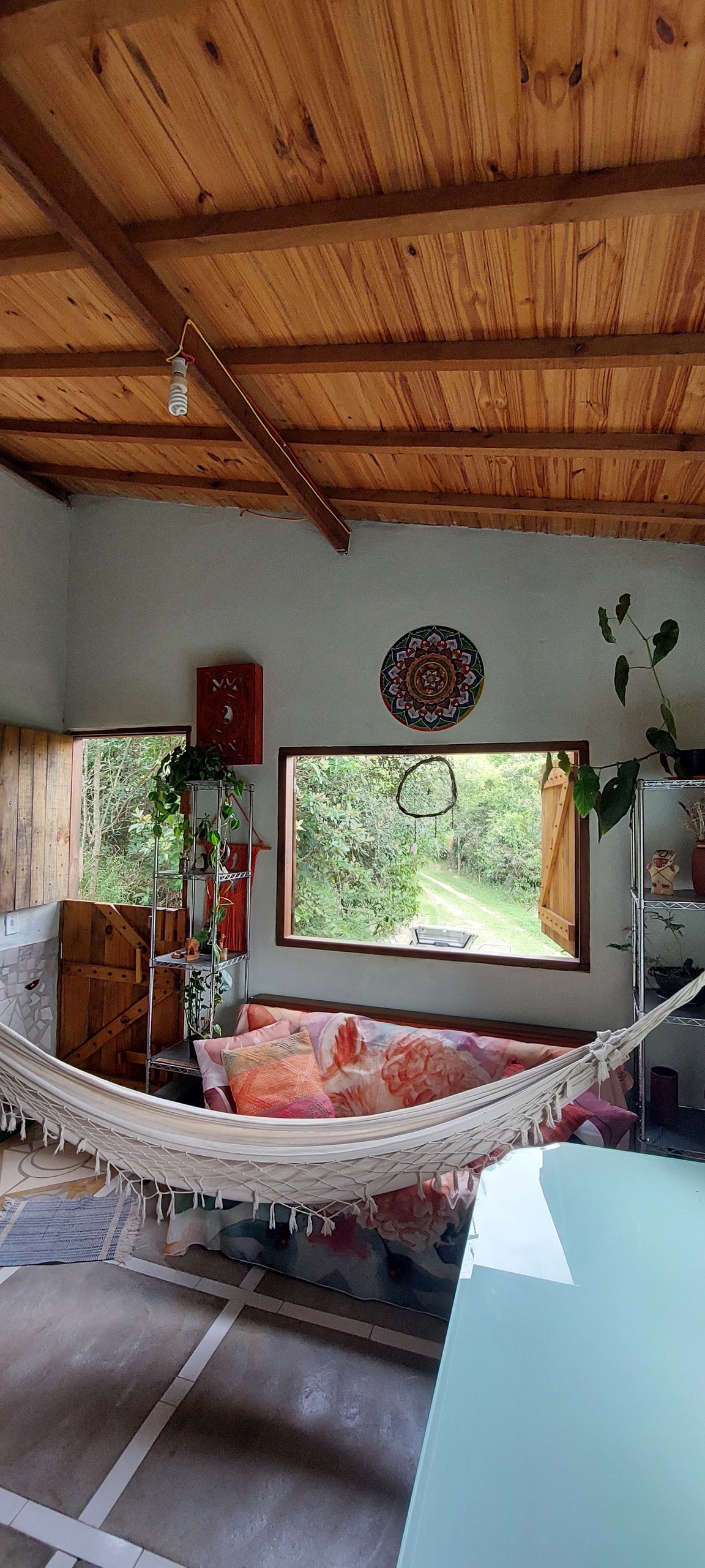 A cozy living space features a spacious hammock hanging between wooden beams, providing a place to relax. A large window offers views of the surrounding greenery, while colorful cushions adorn a small sofa. Potted plants add a touch of nature indoors.
